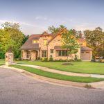 Elegant stone family home with manicured lawn, trees, and driveway on a sunny day.
